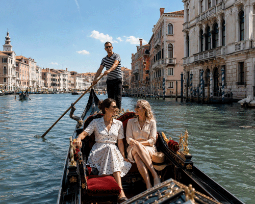 Two women relaxing in a gondola on a sunlit Venetian canal, passing ornate palaces and a striped-shirt gondolier — classic Venice gondola ride with High Point Holidays.