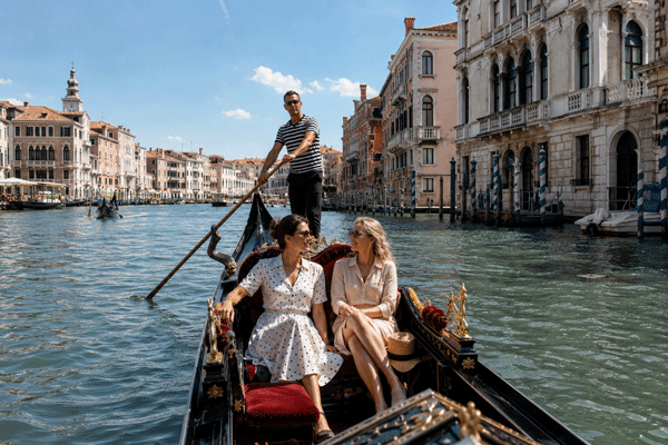 Two women relaxing in a gondola on a sunlit Venetian canal, passing ornate palaces and a striped-shirt gondolier — classic Venice gondola ride with High Point Holidays.