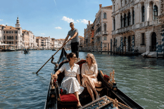 Two women relaxing in a gondola on a sunlit Venetian canal, passing ornate palaces and a striped-shirt gondolier — classic Venice gondola ride with High Point Holidays.